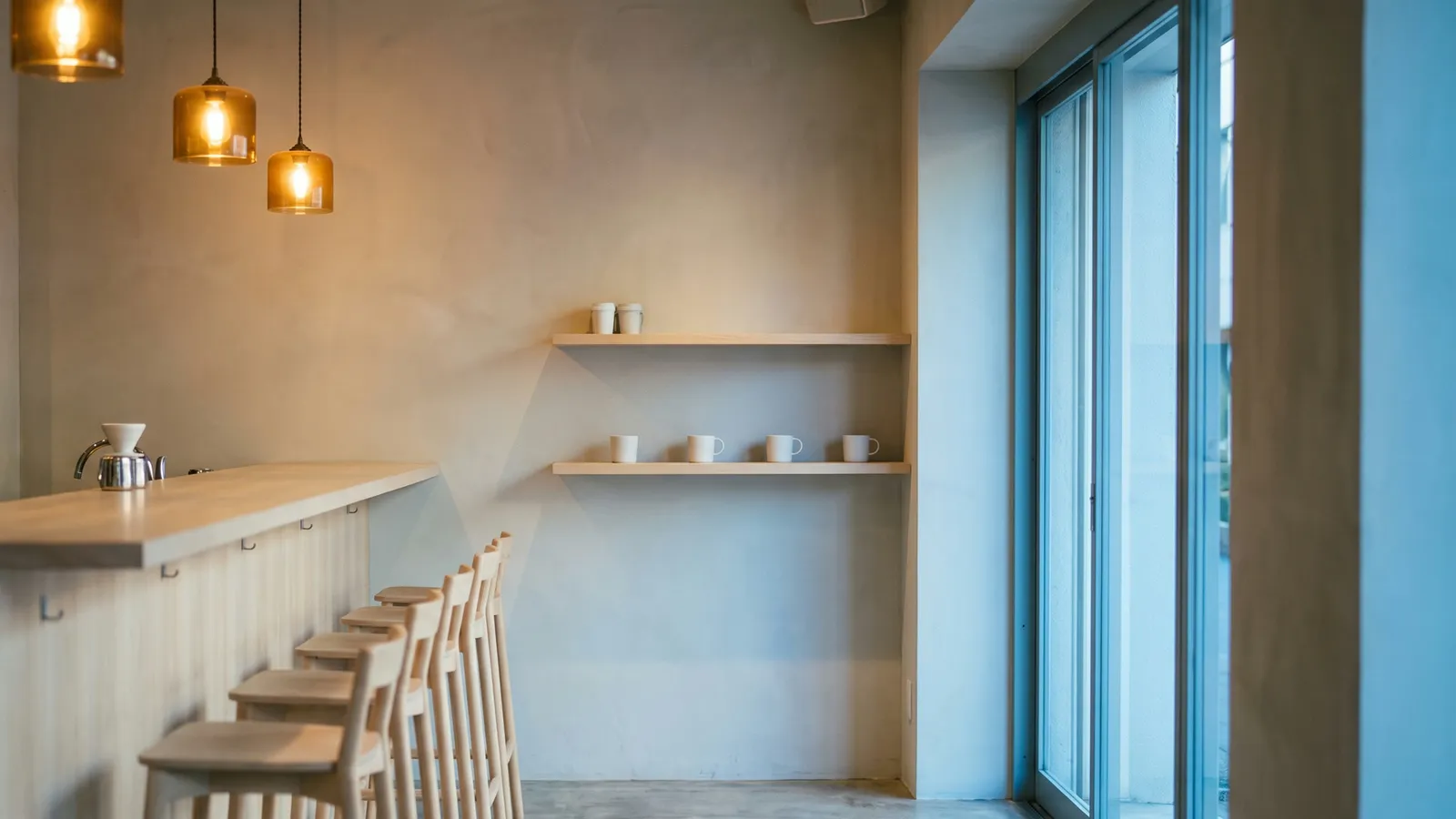Interior del café: mesón de madera clara, piso de cemento pulido y lámparas ámbar sobre la barra.